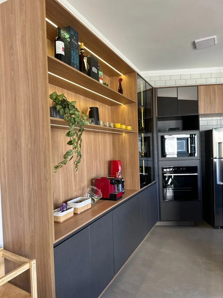 Bar planejado em madeira com nichos iluminados e cozinha com armários pretos e forno embutido ao fundo