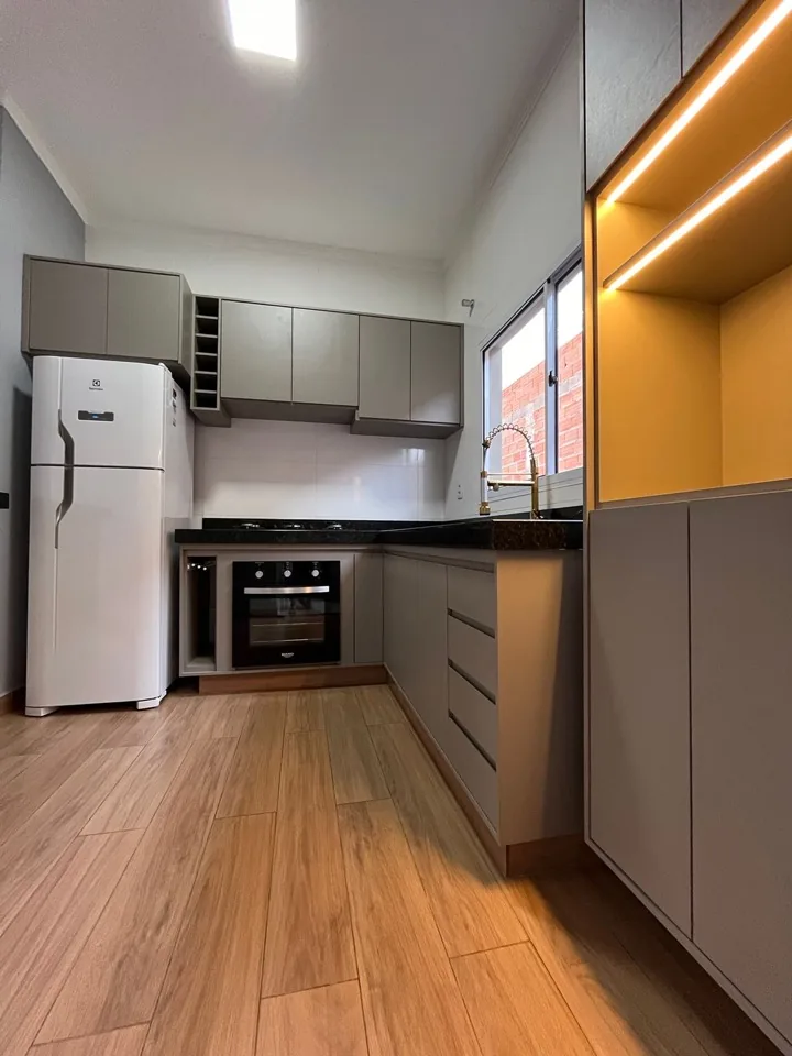 Cozinha compacta em MDF fendi com nicho iluminado em amarelo, piso amadeirado e bancada de granito preto