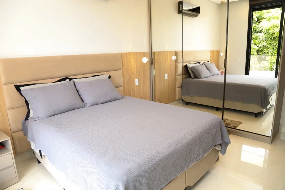 Suíte de casal ampla com cabeceira estofada bege em módulos horizontais, painel de madeira e guarda-roupa de portas espelhadas que refletem o quarto e a vegetação da janela