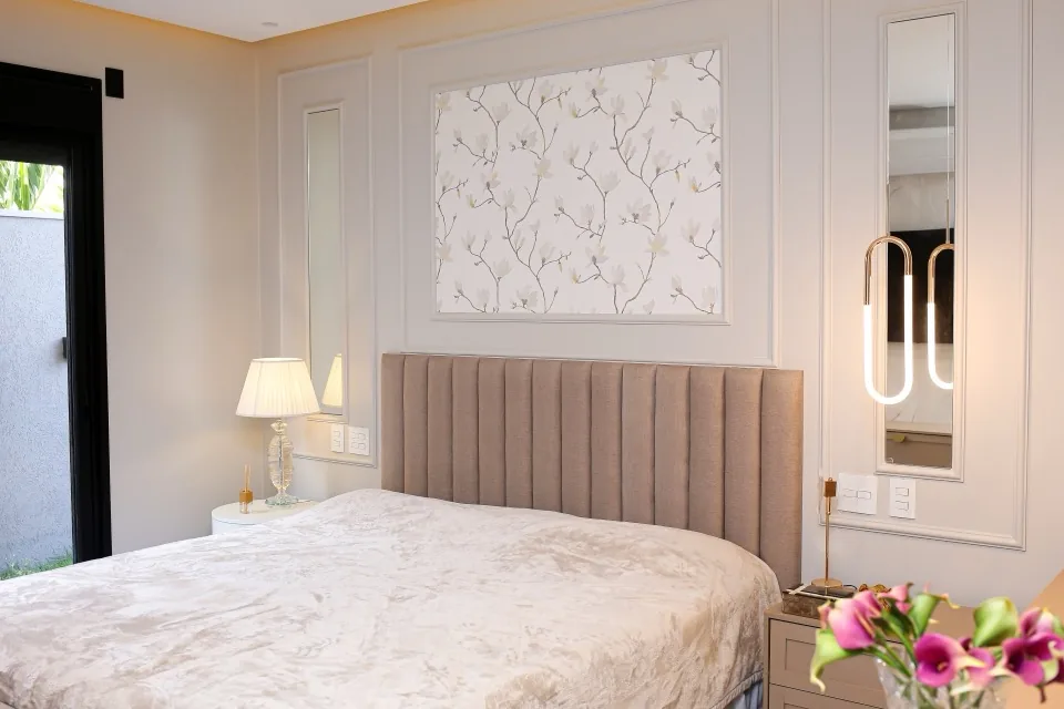 Quarto de casal em estilo provençal com cabeceira canelada bege, boiserie branca na parede, papel de parede floral emoldurado e espelhos verticais com pendente dourado