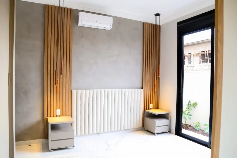 Parede de cabeceira com painel estofado branco canelado entre colunas de ripas de madeira, parede de cimento queimado, pendentes de cobre e piso porcelanato marmorizado